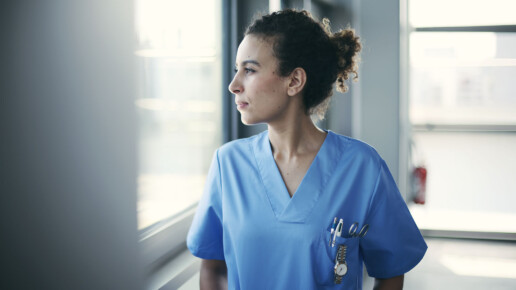 A young female healthcare worker wearing blue medical scrubs stands near a window, gazing outside with a thoughtful expression. She has curly hair tied back and has a watch and pens clipped to her scrub pocket. Natural light streams in through the window, creating a soft, reflective mood.