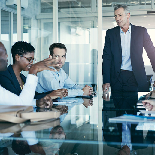 Diverse group of business professionals engaged in a leadership meeting, with a senior executive standing and leading the discussion in a modern conference room.