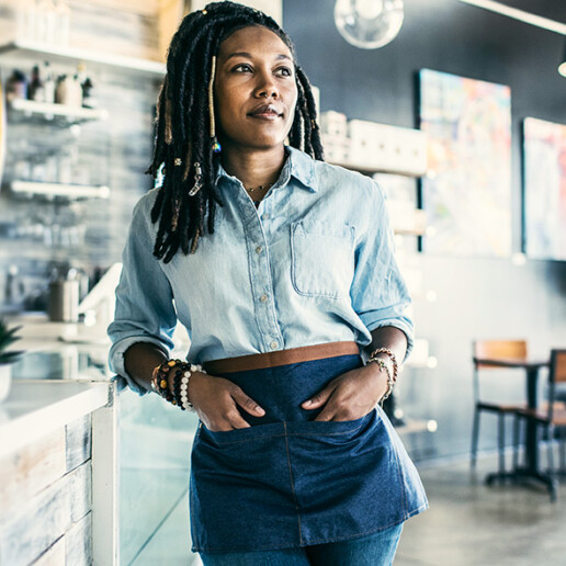 Confident café owner in a denim shirt and apron stands behind the counter in a modern coffee shop, looking thoughtfully into the distance.