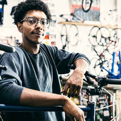 A confident young bicycle mechanic with glasses and curly hair stands in a well-stocked bike shop, leaning on a bike with tools and equipment in the background.
