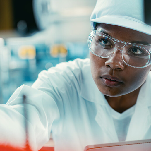 A focused female scientist or technician in a white lab coat, safety glasses, and a cap working with precision in a laboratory or manufacturing environment.