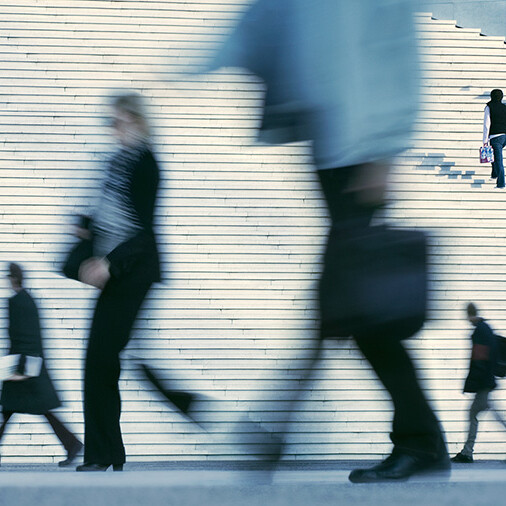 Blurred motion of business professionals walking past a large staircase, symbolizing fast-paced work environments, career progression, and urban life.