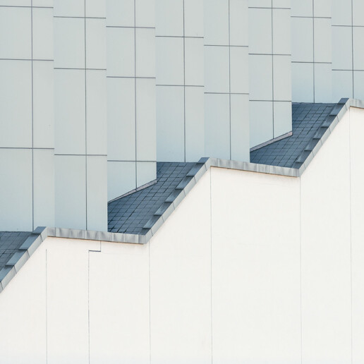 A minimalist architectural facade featuring a series of repeating geometric roof patterns with clean white and gray surfaces, creating a stepped visual effect.