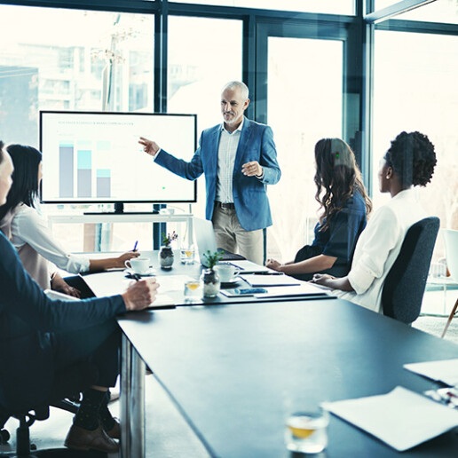 Business professional presenting data on a screen to a diverse group of colleagues in a modern conference room with floor-to-ceiling windows, representing corporate strategy and leadership discussions.