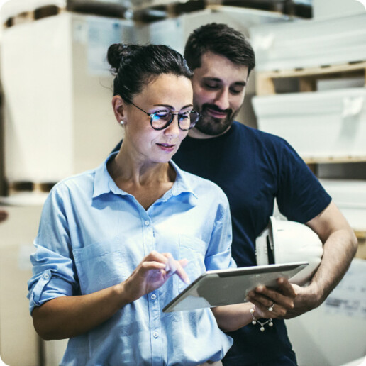 Two professionals in a warehouse setting reviewing information on a tablet. The woman in a blue shirt is interacting with the device, while the man in a dark shirt holds a hard hat and looks on.