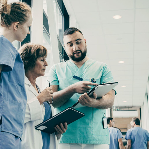 Healthcare professionals in a hospital corridor discussing patient care, with one nurse pointing at a clipboard while colleagues listen attentively.