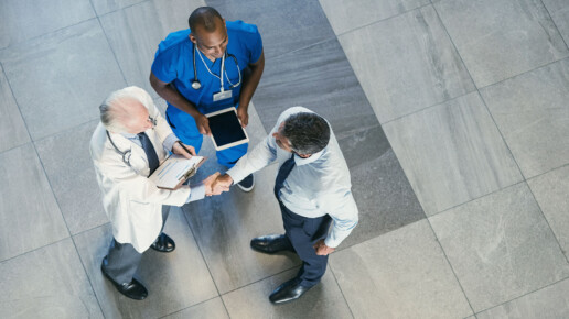 Overhead view of two doctors, one in a white coat and one in blue scrubs, meeting with a business professional and shaking hands in a hospital or clinic setting.