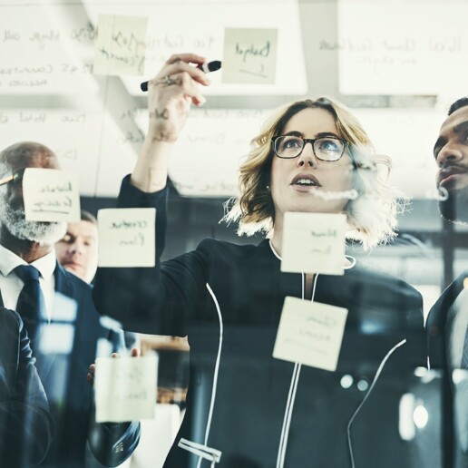 Business professionals collaborating in front of a glass wall covered with sticky notes and handwritten strategy ideas, with one woman leading the discussion and pointing to the board.