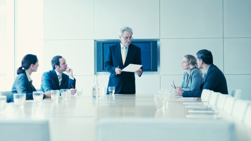 A senior executive stands at the head of a modern conference table, holding a document and addressing four colleagues seated around the table in a corporate boardroom. The group appears engaged in a formal business meeting, with glasses of water and notebooks on the table.