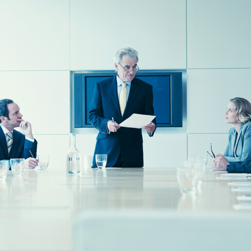 A senior executive stands at the head of a modern conference table, holding a document and addressing four colleagues seated around the table in a corporate boardroom. The group appears engaged in a formal business meeting, with glasses of water and notebooks on the table.