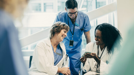 Three health care professionals in scrubs and lab coats smiling and looking at a mobile device in a bright hospital environment.