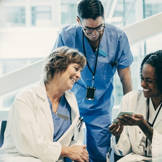 Three health care professionals in scrubs and lab coats smiling and looking at a mobile device in a bright hospital environment.