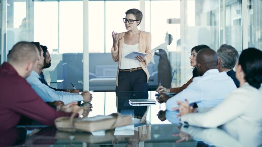 A businesswoman stands at the head of a conference table presenting to a diverse group of colleagues, holding a tablet and speaking confidently during a meeting in a modern glass-walled office.