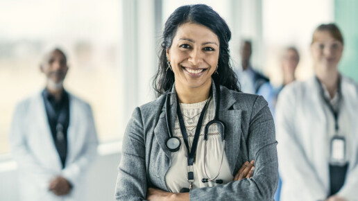 A woman health care leader wearing a stethoscope stands confidently with a clinical team blurred behind her.