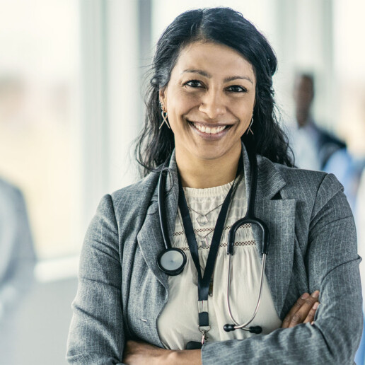 A woman health care leader wearing a stethoscope stands confidently with a clinical team blurred behind her.