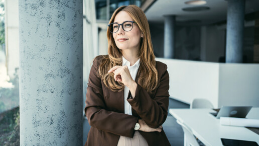 Professional woman standing in a modern office space, looking out thoughtfully.